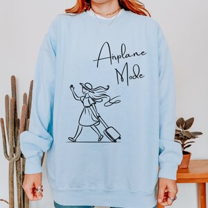Pu&ograve; includere: Felpa blu chiaro con una grafica nera di una donna con un cappello che tira una valigia con il testo "Airplane Mode" su di essa.