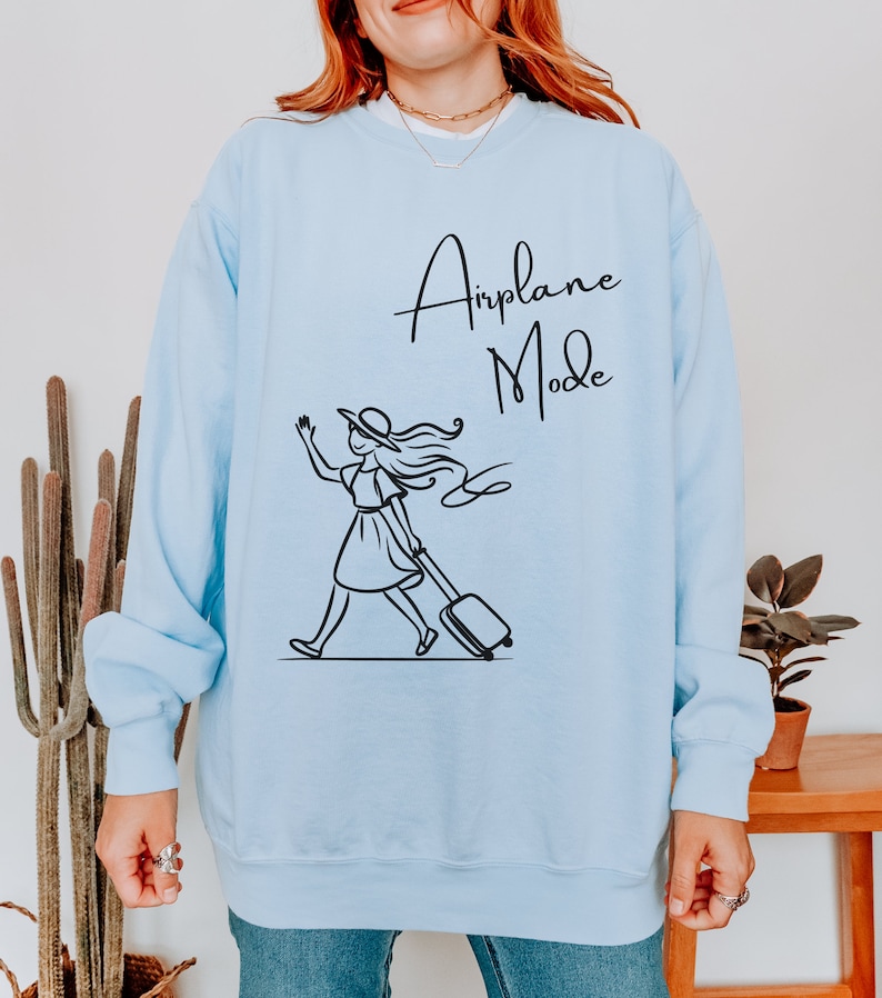 Pu&ograve; includere: Felpa blu chiaro con una grafica nera di una donna con un cappello che tira una valigia con il testo "Airplane Mode" su di essa.