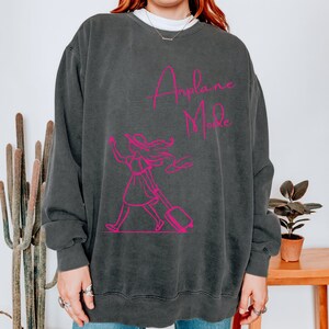 May include: A dark gray sweatshirt with a pink graphic of a woman walking with a suitcase and the text "Airplane Mode".