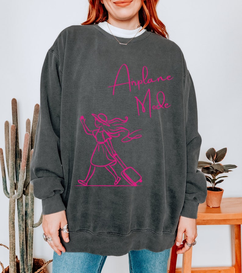 May include: A dark gray sweatshirt with a pink graphic of a woman walking with a suitcase and the text "Airplane Mode".