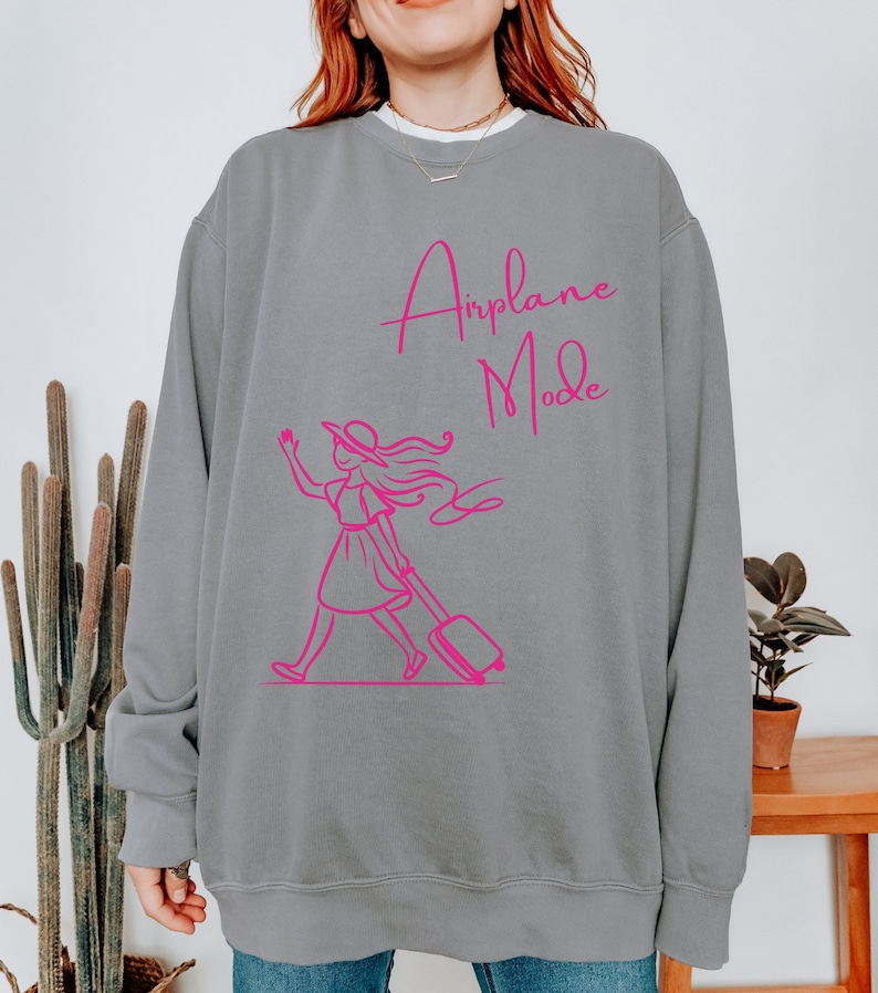 May include: Gray sweatshirt with a pink graphic of a woman pulling a suitcase and the text "Airplane Mode".