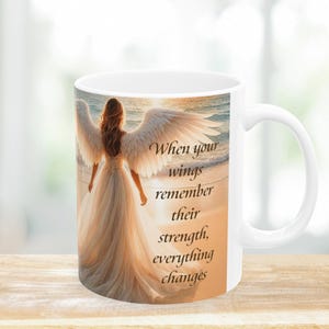 Pode incluir: Caneca de cer&acirc;mica branca com um anjo com grandes asas brancas em uma praia ao p&ocirc;r do sol. A caneca tem o texto "When your wings remember their strength, everything changes" impresso.