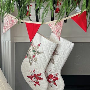 以下が含まれることがあります： 星型のデザインが施された、2つの白いキルティングクリスマスストッキング。ストッキングは、赤と白の花柄のパッチワークスターデザインです。緑と赤と白のペナントフラッグのガーランドが上に吊り下げられています。
