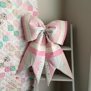 May include: A large patchwork bow in pink, white, and mint green hues, displayed on a white ladder. The bow's fabric is patterned with floral and geometric designs. A matching patchwork quilt, also with floral and geometric patterns, is visible in the background.
