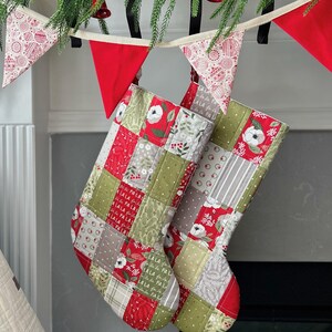 Puede incluir: Dos calcetines navideños acolchados con patrones de patchwork en rojo, verde y gris. Los calcetines están decorados con diseños florales y geométricos. Una guirnalda roja y blanca y vegetación cuelgan encima.