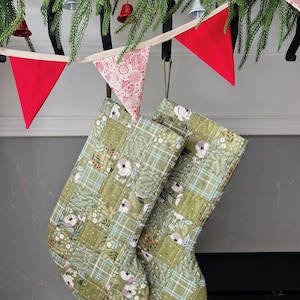 以下が含まれることがあります： 緑色の花柄とチェック柄のキルティングクリスマスストッキング2つが、緑と赤いペナントフラッグのガーランドの下に吊るされています。ストッキングは、白い花と赤いベリーのパッチワークデザインです。