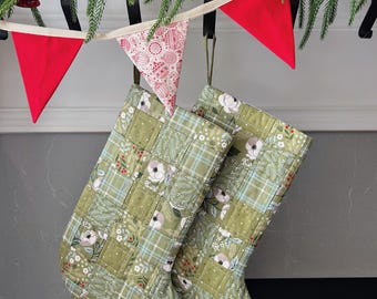 May include: Two quilted Christmas stockings with a green floral and plaid pattern hang below a garland of greenery and red pennant flags. The stockings have a patchwork design with white flowers and red berries.