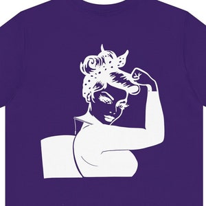 May include: Purple t-shirt with a white silhouette of a woman with her fist raised in the air. She has a polka dot bandana in her hair.