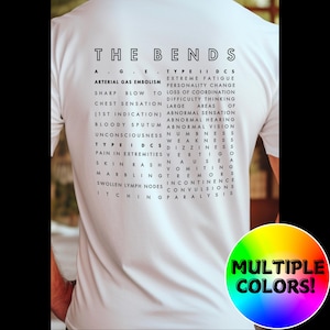 May include: White t-shirt with black text that reads "THE BENDS" and lists symptoms of decompression sickness, also known as "the bends". The text is in a word search format. The bottom of the shirt has the text "MULTIPLE COLORS!" in a rainbow of colors.