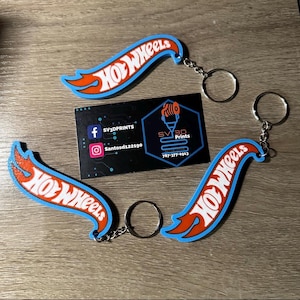 Puede incluir: Dos llaveros Hot Wheels azules y naranjas con anillos de llaves plateados. Los llaveros tienen la forma del logotipo de Hot Wheels. Los llaveros están sobre una superficie de madera con una tarjeta de visita para SV3D Prints.