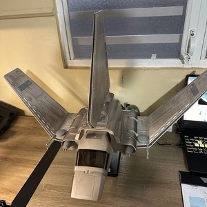 Puede incluir: Un modelo de caza TIE Interceptor de Star Wars gris y blanco con una cabina y alas detalladas. El modelo está en un soporte negro.