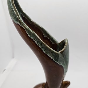 Puede incluir: Un jarrón de cerámica marrón con un esmalte verde. El jarrón tiene forma de cuerno curvo y una abertura ancha en la parte superior.