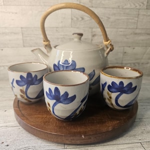 May include: A ceramic tea set with a teapot and four cups. The set features a blue and white floral design on a light gray background. The teapot has a bamboo handle and the cups are sitting on a wooden tray.