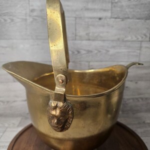 Puede incluir: Un cubo de hielo de latón dorado con un asa y una cabeza de león decorativa en el lateral. El cubo está sobre una superficie de madera.