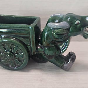 May include: A ceramic planter in the shape of a green ox pulling a cart. The ox has gray horns and a yellow nose. The cart has a rectangular opening for plants and a decorative wheel.
