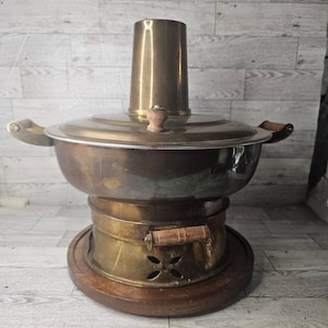 May include: A vintage brass hot pot with a central chimney, lid, and wooden handles. The pot sits on a wooden base with a brass burner section featuring decorative cutouts. The design suggests a traditional cooking vessel.