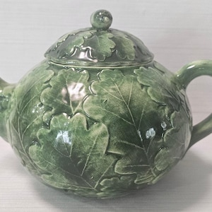 May include: A green ceramic teapot with a lid, spout, and handle. The teapot is decorated with a leaf pattern. The teapot is approximately 8 inches in height and 10 inches in width.