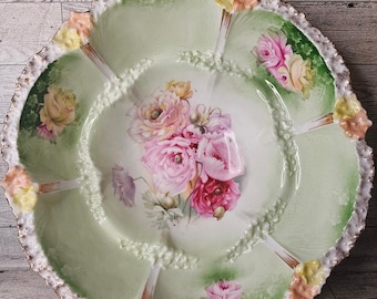 May include: A vintage green and white porcelain bowl with a floral design. The bowl has a scalloped edge and is decorated with pink, yellow, and red roses. The bowl is perfect for serving fruit, snacks, or desserts.
