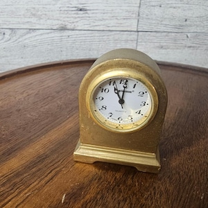 Könnte beinhalten: Eine kleine, goldfarbene Quarzuhr mit weißem Zifferblatt und schwarzen Ziffern. Die Uhr hat eine abgerundete Oberseite und einen rechteckigen Sockel. Das Zifferblatt zeigt "Catium" und "QUARTZ".