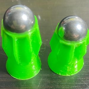 Pode incluir: Dois lançadores de brinquedo de plástico verde com bolas de metal prateadas dentro. Os lançadores têm a forma de um cone com a parte superior arredondada.