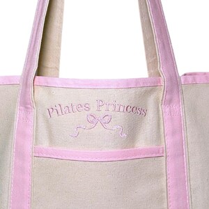 May include: A light pink canvas tote bag with pink handles and a pink trim. The bag has a front pocket and is embroidered with the text "Pilates Princess" in pink thread.