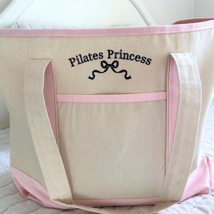 May include: Large pink canvas tote bag with black embroidered text that says "Pilates Princess" with a ribbon design. The bag has a pink trim and a large pocket on the front.