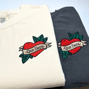 Peut inclure: Deux t-shirts avec des cœurs rouges brodés et des feuilles vertes. Le texte sur la bannière dit "Que Sera."