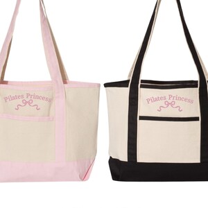 May include: Two canvas tote bags with black and pink trim. The bags have a front pocket and the text "Pilates Princess" embroidered in pink on the front.