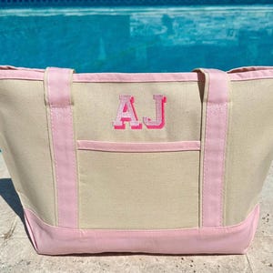 May include: A beige canvas tote bag with pink trim and handles. The bag has a large front pocket and the initials "AJ" embroidered in pink on the front.