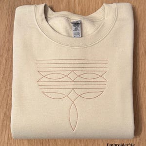 May include: A light brown sweatshirt with a geometric design embroidered on the front in a light brown thread. The design features a series of curved lines and circles. The sweatshirt is folded in half and laid on a wooden surface. The text "EmbroiderMe Studio" is visible in the bottom right corner of the image.