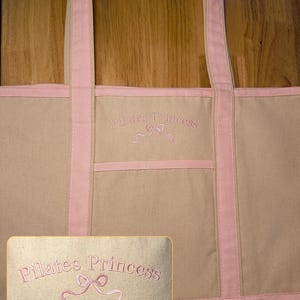 May include: A large pink canvas tote bag with pink straps and a pink embroidered design that says "Pilates Princess" with a pink ribbon bow.
