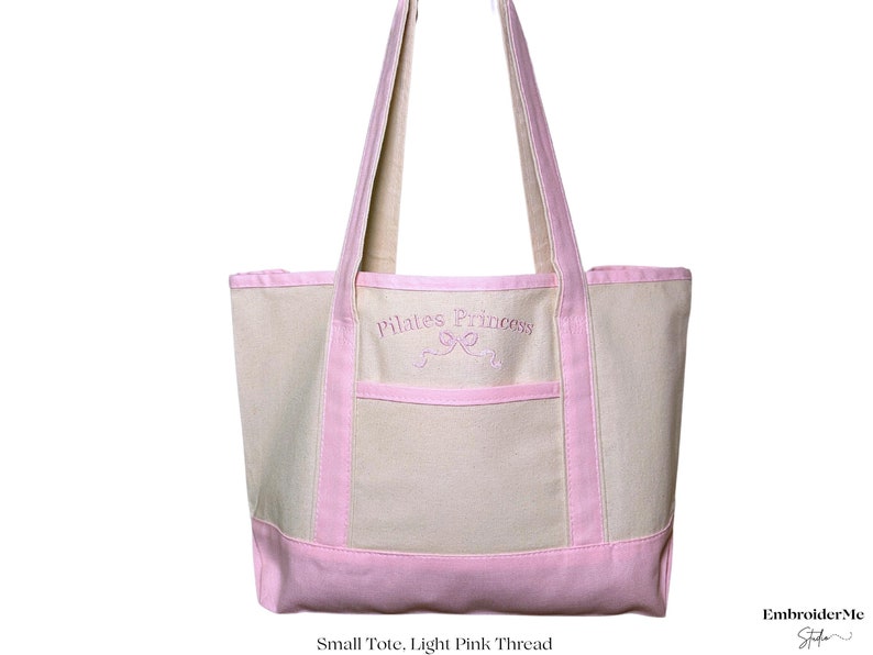 May include: A light pink canvas tote bag with pink handles and a pink trim. The bag has a front pocket and is embroidered with the text "Pilates Princess" in pink thread.