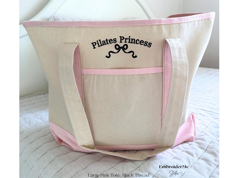 May include: Large pink canvas tote bag with black embroidered text that says "Pilates Princess" with a ribbon design. The bag has a pink trim and a large pocket on the front.
