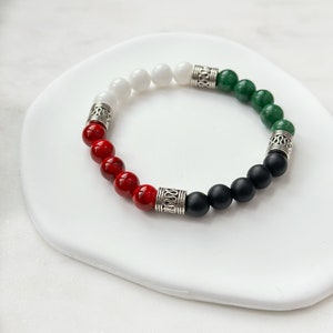 May include: A bracelet with red, white, black, and green beads separated by silver spacers. The bracelet is on a white surface.