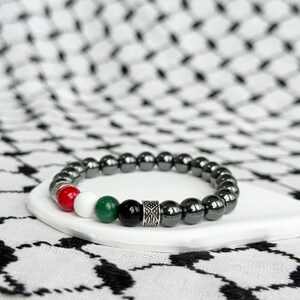 May include: A black hematite beaded bracelet with red, white, green, and black accent beads and a silver charm. The bracelet is on a white surface.