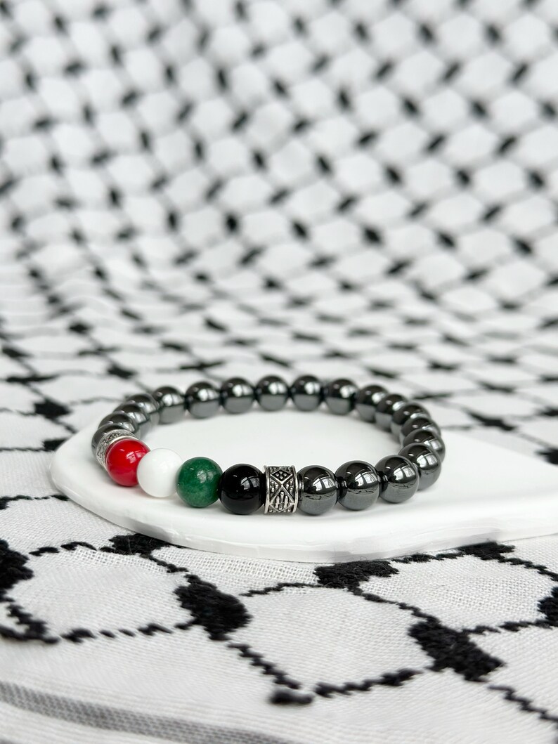 May include: A black hematite beaded bracelet with red, white, green, and black accent beads and a silver charm. The bracelet is on a white surface.