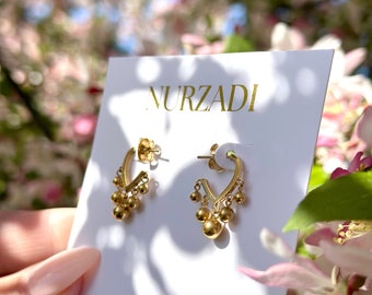 May include: Gold hoop earrings with a dangling chain of small gold balls. The earrings are displayed on a white card with the word "NURZADI" printed in gold lettering.