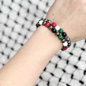 May include: Two beaded bracelets on a wrist. The bracelets are made of black, white, red, and green beads. The beads are interspersed with silver spacers.