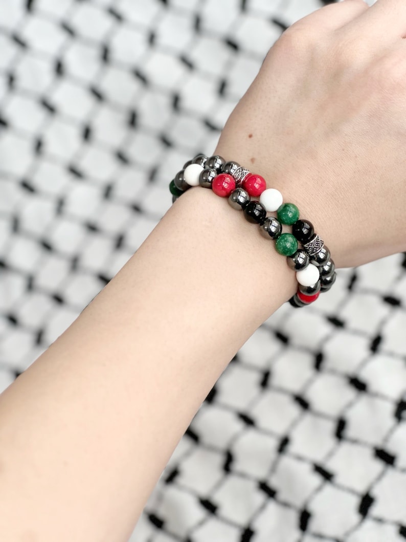 May include: Two beaded bracelets on a wrist. The bracelets are made of black, white, red, and green beads. The beads are interspersed with silver spacers.