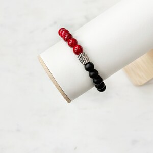 May include: A red and black beaded bracelet with a silver charm. The bracelet is displayed on a white stand.