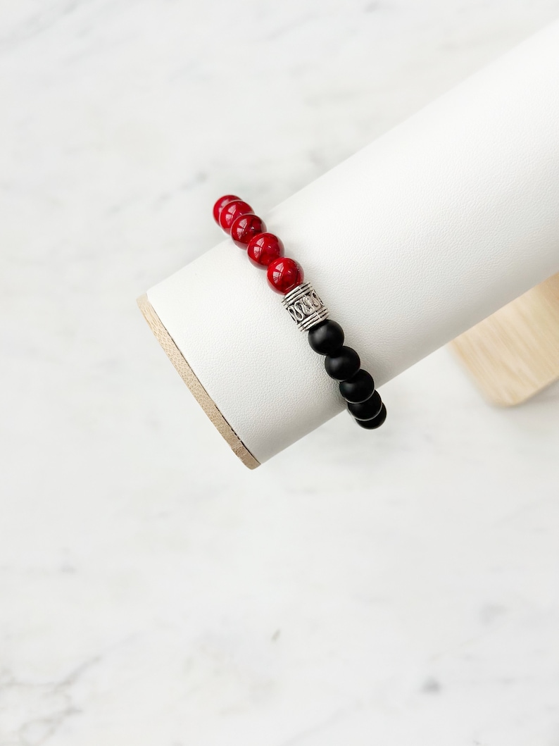 May include: A red and black beaded bracelet with a silver charm. The bracelet is displayed on a white stand.