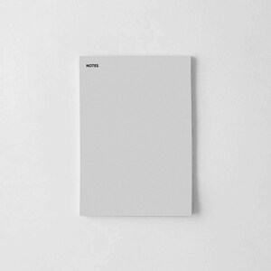 Puede incluir: Un bloc de notas rectangular gris claro con la palabra "NOTES" impresa en negro en la esquina superior izquierda. El bloc de notas está sobre un fondo blanco, creando una estética minimalista. El bloc de notas está en blanco, listo para usar.