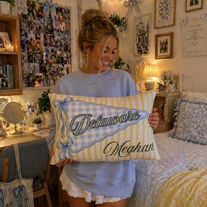 May include: A rectangular cushion with a blue and yellow striped and checkered design. The cushion has the word "Delaware" in script and the name "Meghan" in a matching script. The cushion has blue bow accents.