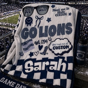 May include: A white and navy blue custom blanket with the text "GO LIONS" and "Sarah". The blanket has a checkered border and various graphics, including sunglasses that say "WE win". A navy blue water bottle with "CUSTOM FOOTBALL" is also visible.