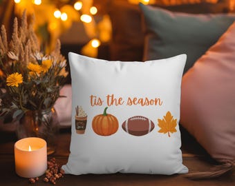 May include: White square pillow with the text "tis the season" in orange. The pillow features illustrations of a coffee drink, pumpkin, football, and maple leaf. The pillow is on a wooden surface.
