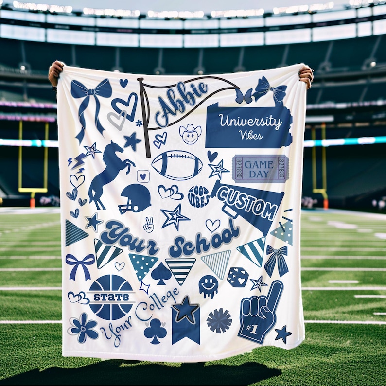 May include: A white blanket with navy blue graphics, including stars, hearts, a football, and the words "Abbie", "University Vibes", "Your School", and "Your College". The blanket is held up in front of a football field.
