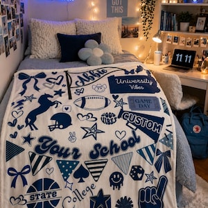 May include: A white and blue custom blanket with various graphics, including a football, stars, and the text "Your School." The blanket also features the name "Abbie" and the phrases "University Vibes" and "Game Day."