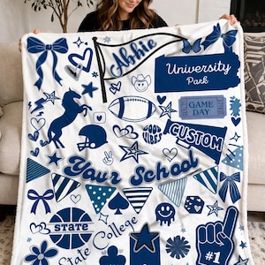 May include: A white fleece blanket with a navy blue and light blue design. The blanket features various graphics, including a football, stars, hearts, bows, and the text "Abbie", "University Park", "Your School", and "State College".