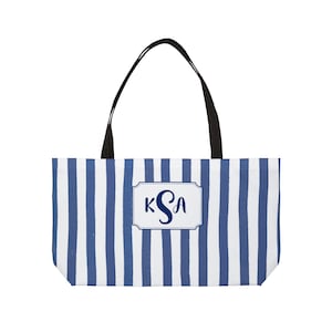 Può includere: Una borsa tote a righe blu e bianche con manici neri. La borsa presenta un'etichetta rettangolare con le iniziali "KSA" in blu navy. La borsa è progettata per trasportare oggetti.