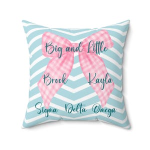 May include: Square pillow with a light blue and white chevron pattern background. A pink gingham bow is centered on the pillow, with the words "Big and Little" above the bow. The names "Brook" and "Kayla" are on the bow's tails, and "Sigma Delta Omega" is below.